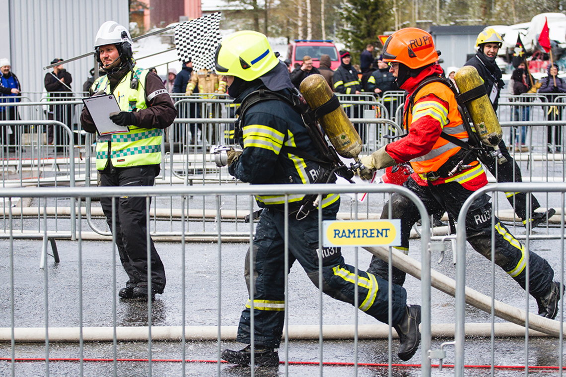Palokuntalaiset juoksevat varusteissaan metallisten aitojen välissä vuoden 2019 palokuntien SM-kilpailussa.