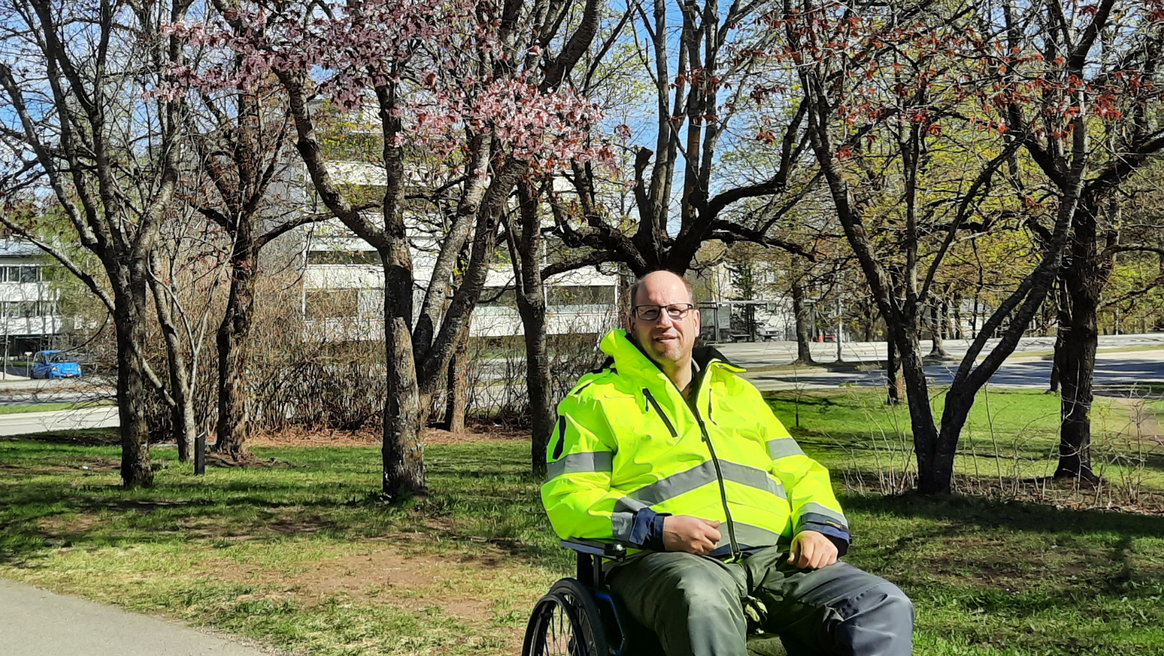 Marko Pousi istuu neonkeltainen työtakki päällä pyörätuolissa. Taustalla kirsikkapuita.