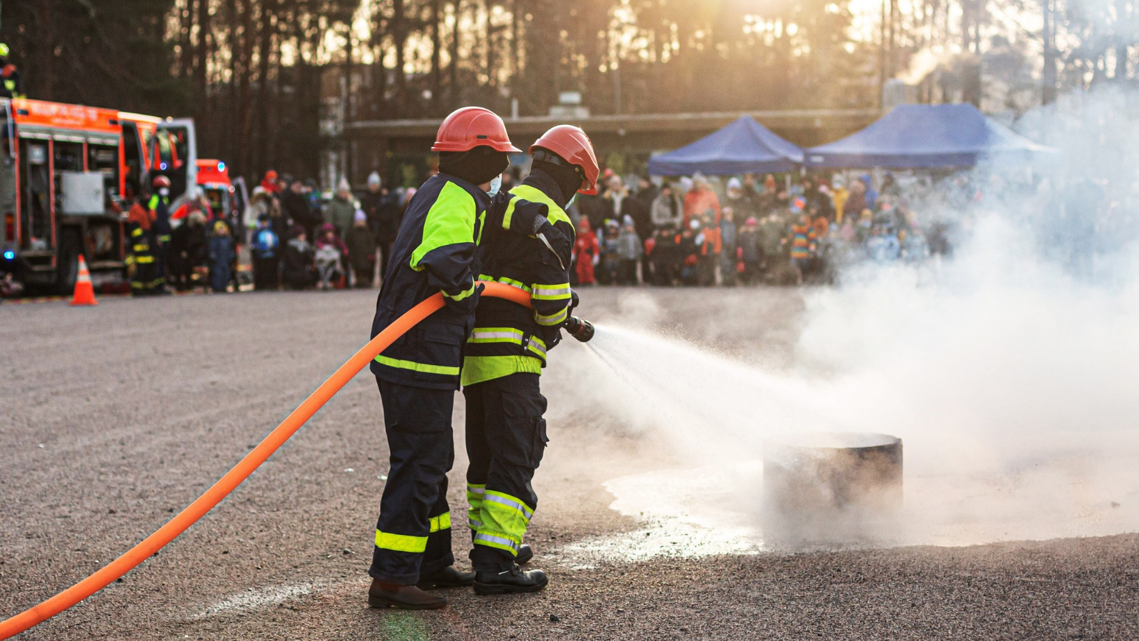 Kuvassa kaksi palokuntanuorta sammuttaa pientä paloa, taustalla näkyy paloauto, telttoja ja ihmisiä, jotka seuraavat sammutustyötä.