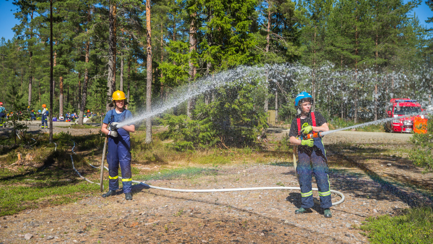 Kaksi palokuntanuorta suihkuttavat letkulla vettä metsäisellä leirialueella.