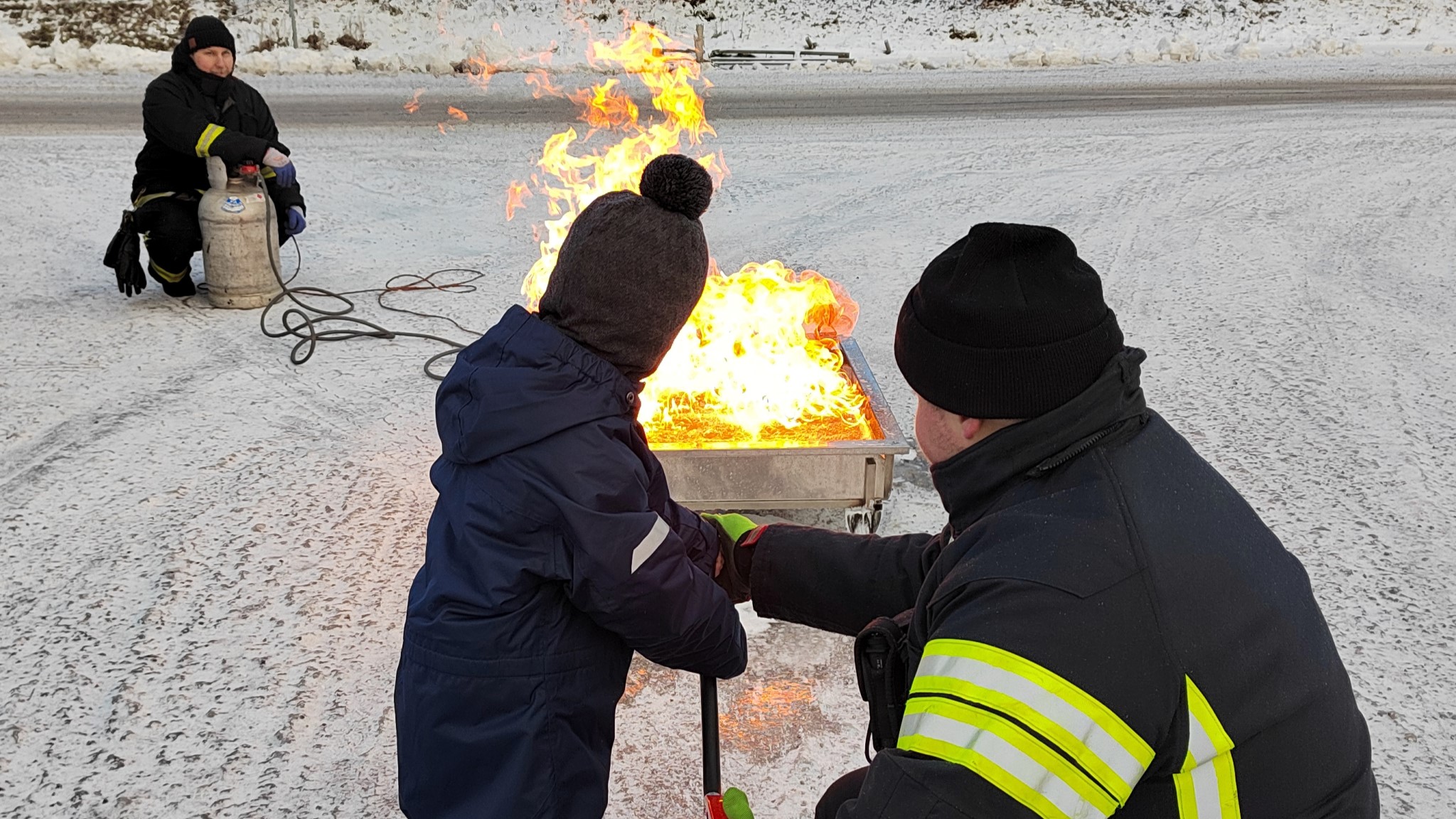 Lapsi osoittaa palosammuttimella liekkejä palokuntalaisen avustuksella.