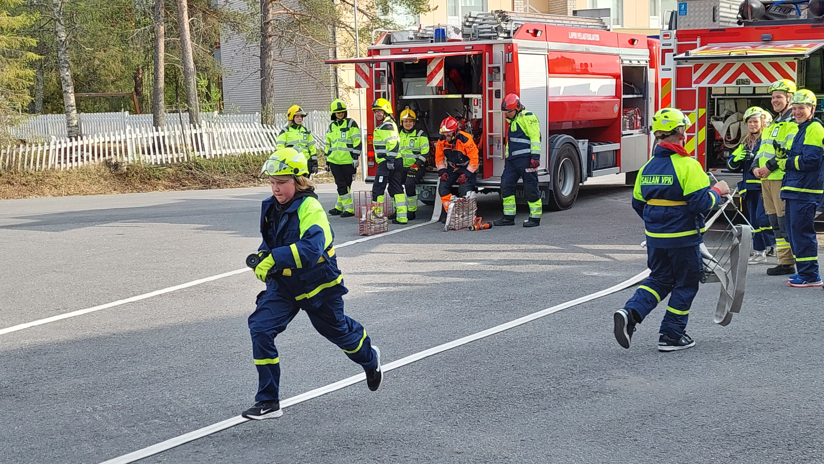 Palokuntanuori juoksee viestikilpailussa samalla, kun hälytysosastolaisten sekä palokuntanuorten joukkueet odottavat paloautojen edessä.