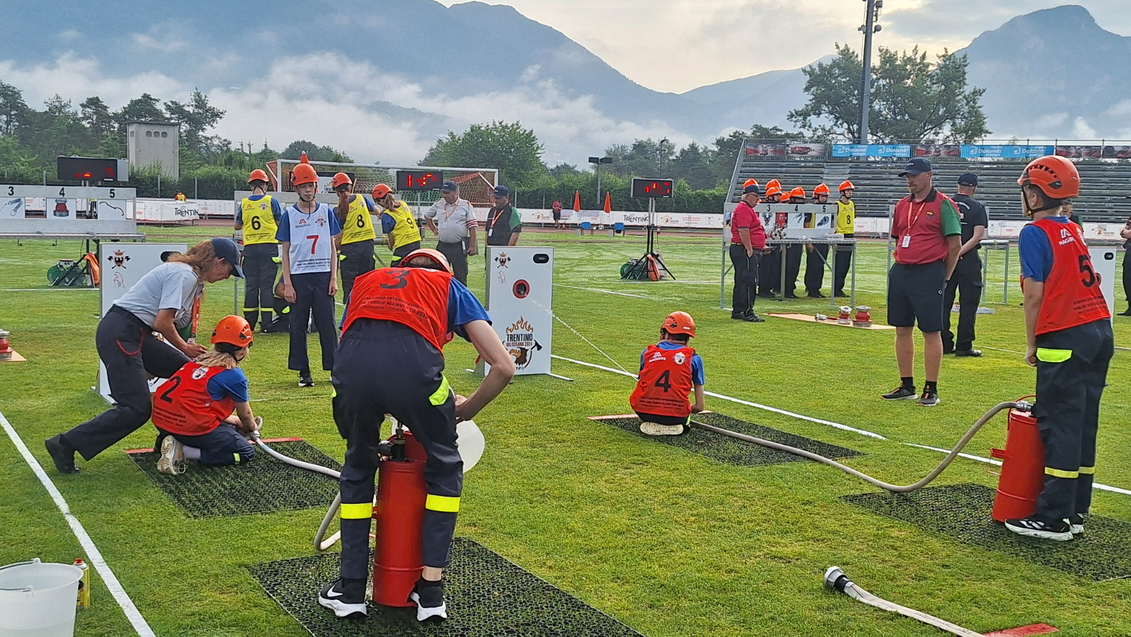 Palokuntanuorten CTIF-kilpailijoita ruohokentällä suorittamassa tehtävää toimitsijoiden tarkkaillessa vierestä. Taustalla vuoret.