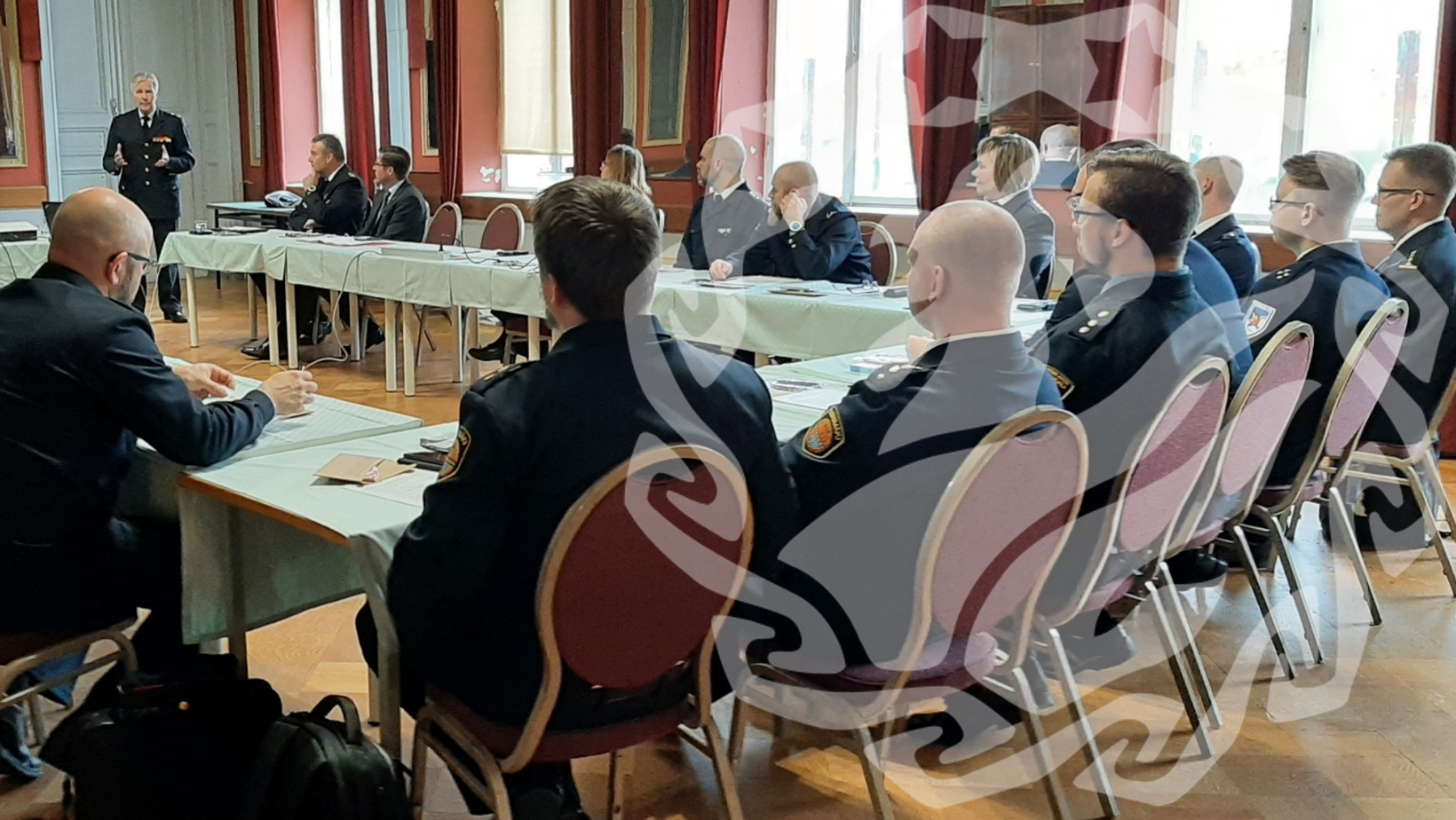 Pitkän pöydän ääressä istuu ihmisiä. Pöydän edessä yksi ihminen puhumassa.