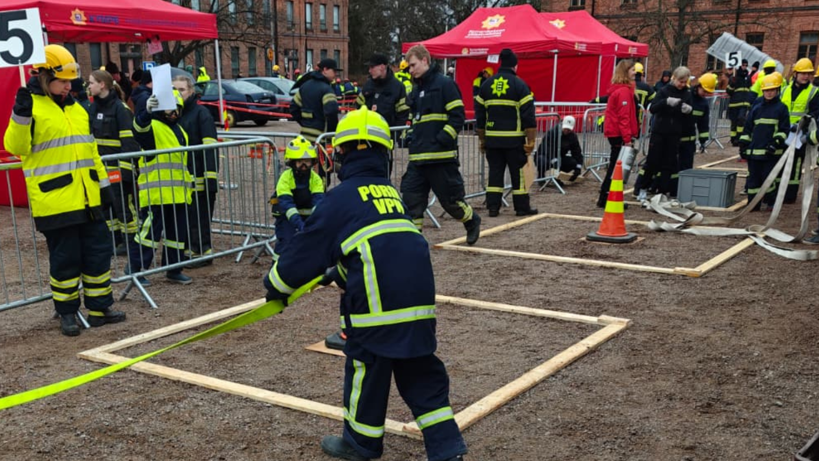 Palokuntanuoria hiekkakentällä suorittamassa kalustokäsittelykilpailun tehtäviä. Kuvassa myös toimitsijoita.